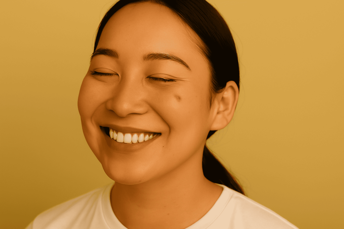 Close-up of a young woman smiling brightly with eyes closed in front of a solid yellow background, showing clear skin and positive expression.