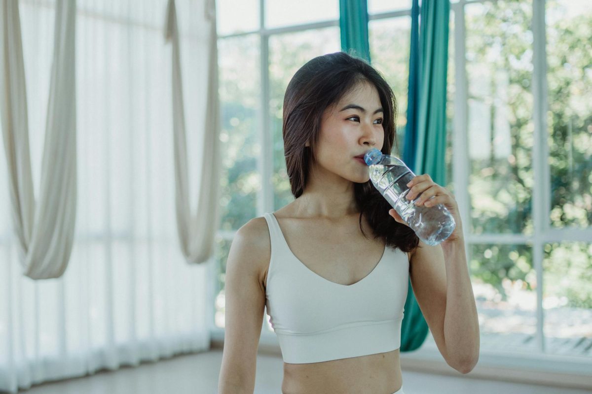 A young Asian woman in sportswear drinking water from a plastic bottle inside a bright studio, with large windows and fitness equipment in the background.