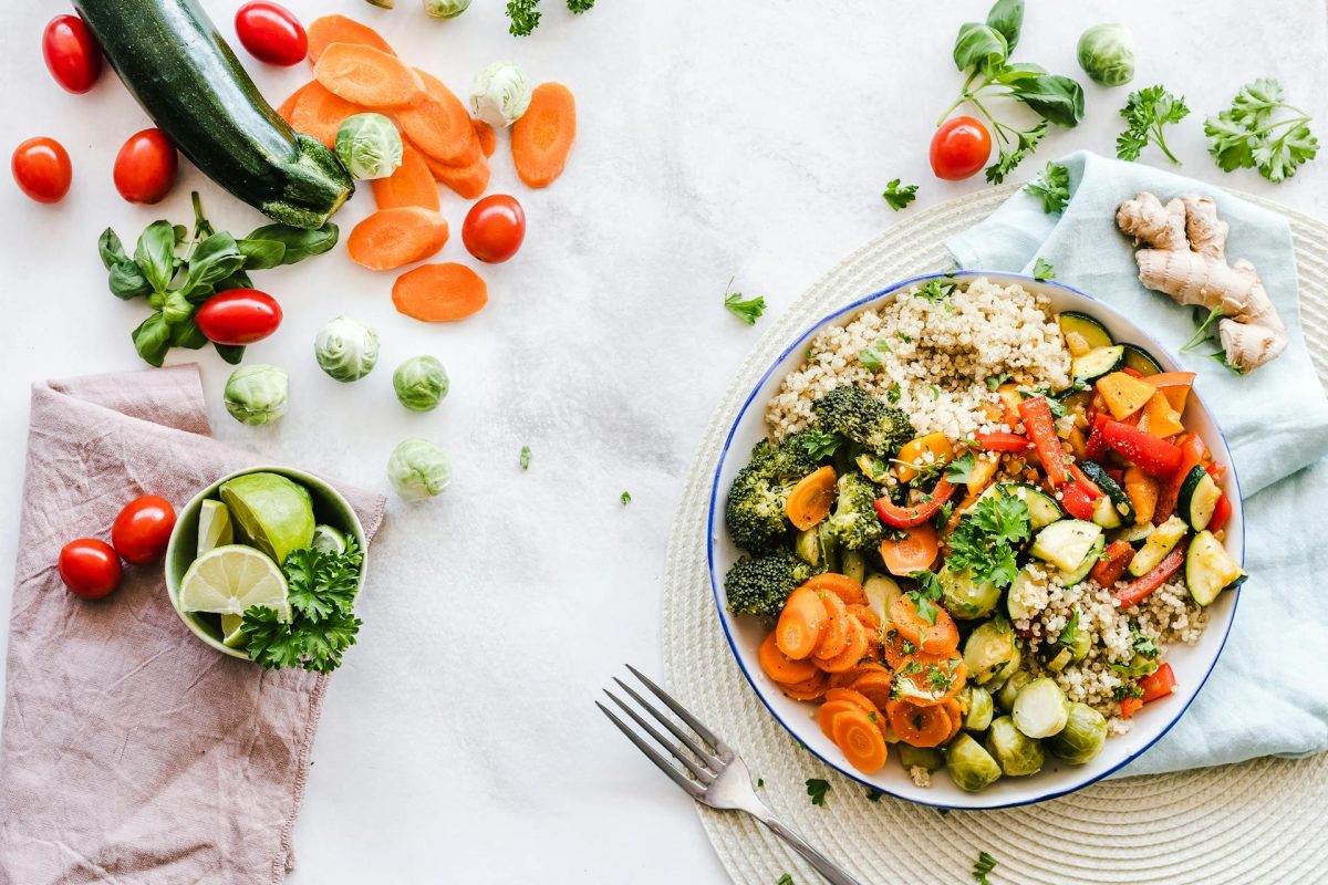 A colourful overhead photo of a balanced plant-based meal featuring cooked vegetables, quinoa, and fresh raw vegetables such as carrots, cherry tomatoes, Brussels sprouts, and lime, arranged on a white background.
