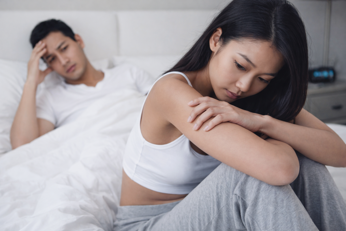 Woman sitting on bed looking distressed while partner appears concerned in the background, illustrating pain during sex.