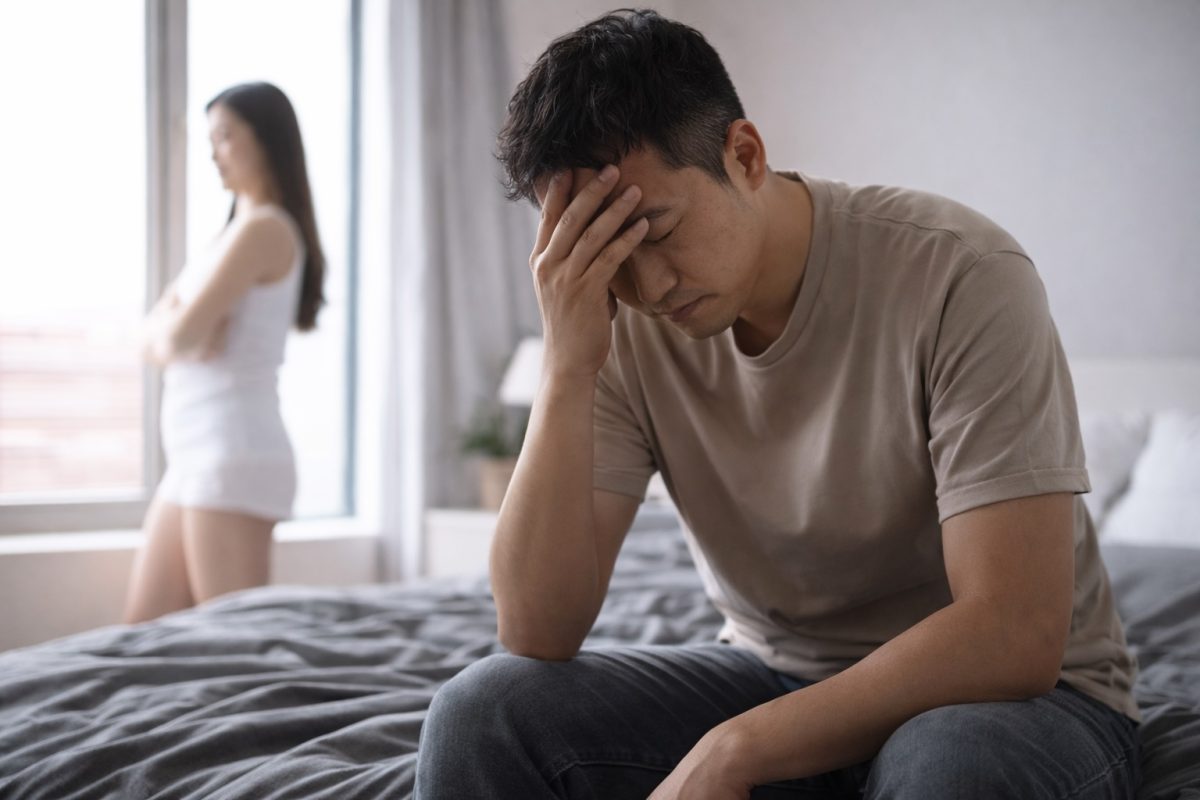 Man sitting on a bed holding his head in distress, representing emotional stress or sexual health concerns affecting relationships
